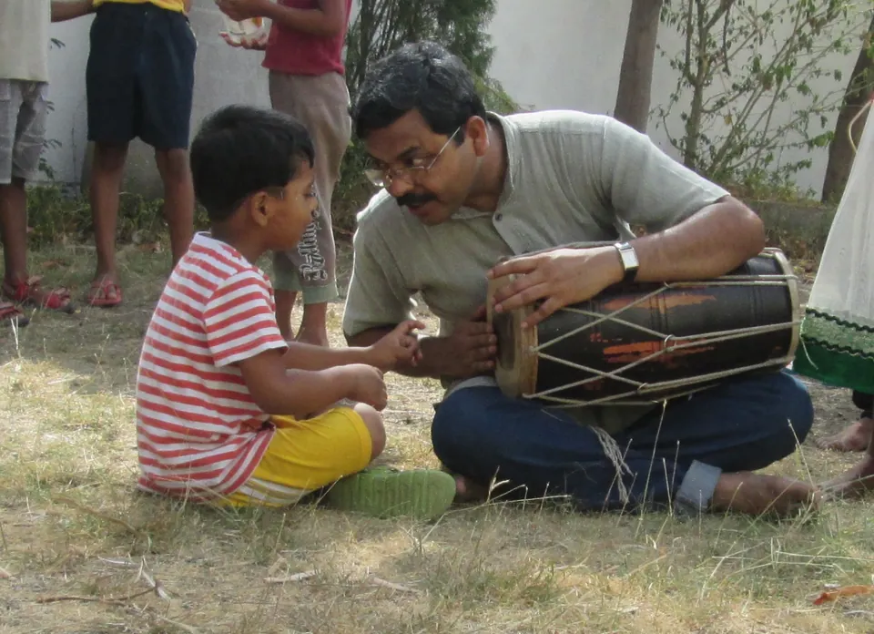 Anil sharing a dholak with a young child while sitting together on the grass